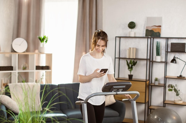 Athlète belle jeune femme concentrée avec un téléphone intelligent en cours d'exécution sur tapis roulant dans la salle de gym