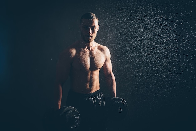 Un athlète blond masculin soulève des haltères avec la main sur une photo de studio de biceps avec des gouttes de pluie