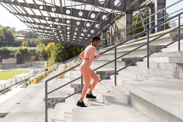 Athlète féminine faisant du fitness dans un survêtement rose près d'un stade de sport, afro-américaine montant les escaliers le matin