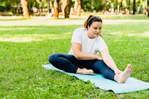 Une athlète féminine grosse et sportive s'étire les jambes sur un tapis de fitness en cours de yoga, s'entraîne à perdre du poids, s'amincit à l'extérieur dans un parc.