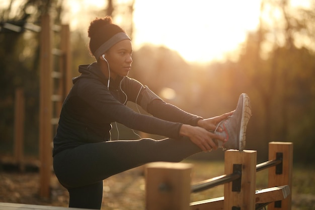 Athlète féminine noire étirant sa jambe tout en faisant de l'exercice dans une salle de sport en plein air au coucher du soleil