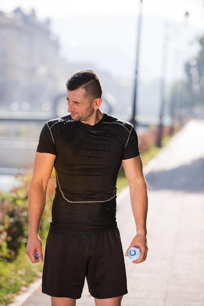 Athlète homme coureur versant de l'eau de la bouteille sur sa tête après avoir fait du jogging dans la ville par une journée ensoleillée