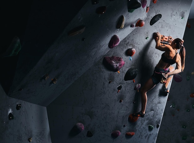 Athlète jeune femme en short et soutien-gorge de sport exerçant sur un mur de bloc à l'intérieur.