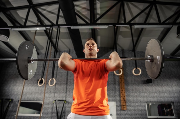 Un athlète masculin portant un t-shirt orange fait un squat avec des poids lourds lors d'une séance d'entraînement