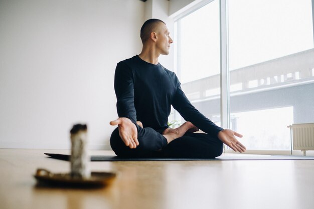 Un athlète masculin en vêtements de sport noirs est assis dans la position du lotus sur fond d'une bougie d'encens dans la salle de sport