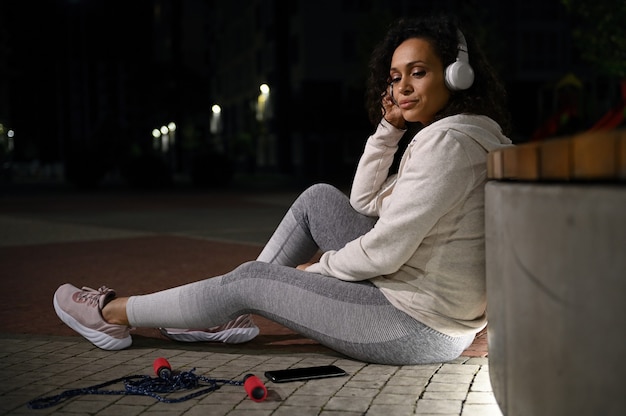 Un athlète de nationalité mixte portant des écouteurs sans fil est assis près d'un banc avec une corde à sauter à proximité. Détente après une séance d'entraînement lors d'une merveilleuse soirée sur le fond illuminé des bâtiments de la ville