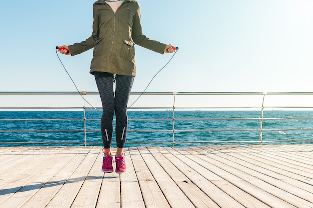 Athlétique femme dans une veste et des baskets saute à la corde le matin sur le fond de la mer