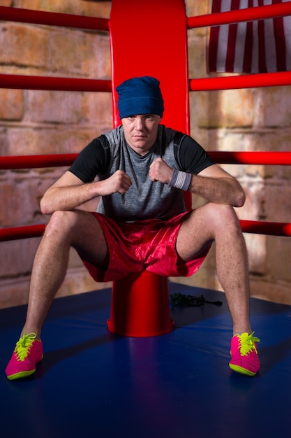 Athlétique jeune boxeur masculin assis près du coin rouge d'un ring de boxe régulier