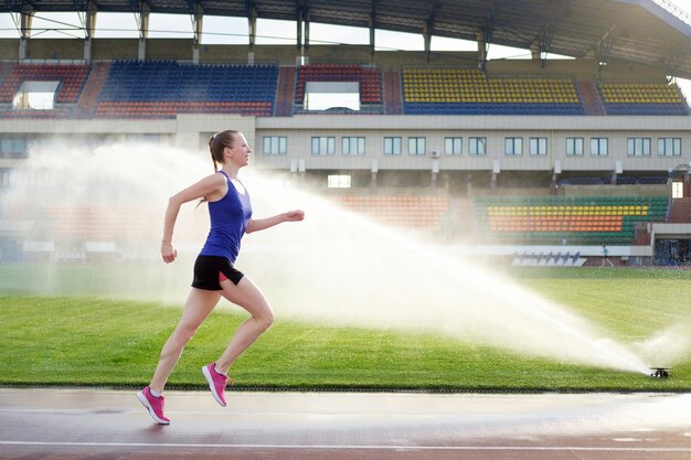 Athlétique jeune femme en baskets roses courir près de l'arroseur sur le stade de la piste de course