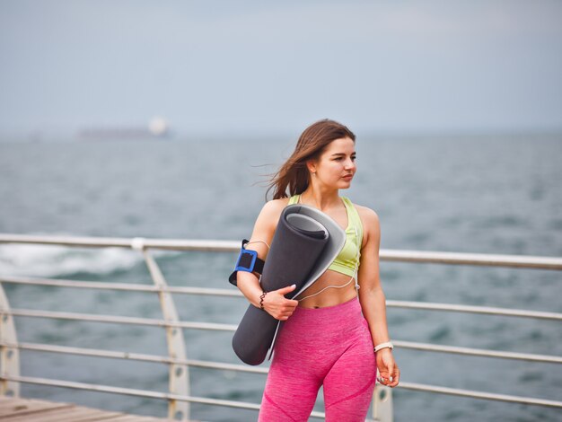 Athlétique Jeune Femme En Tenue De Sport Tient Un Tapis De Yoga Dans Sa Main Et Posant à L'extérieur Sur La Plage