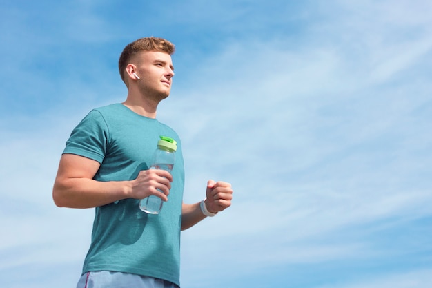 Athlétique mince beau mec coureur jeune bel homme avec une bouteille d'eau écouter de la musique dans les écouteurs sans fil pendant le jogging en cours d'exécution. Copiez l'espace. Concept de mode de vie sain actif. Ciel bleu