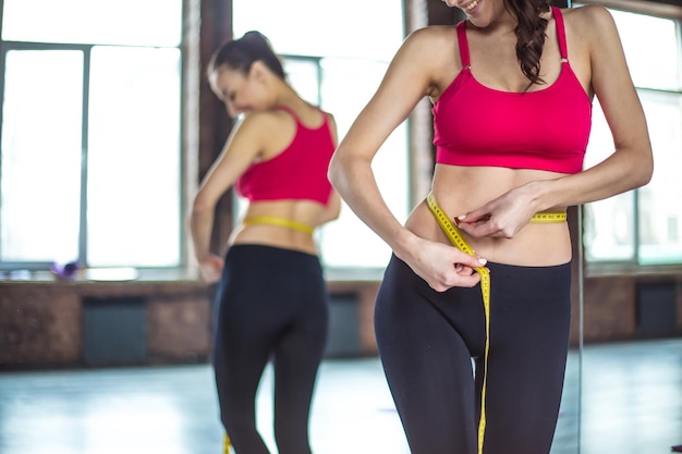 Athlétique souriante femme mince mesurant sa taille par un ruban à mesurer après l'entraînement en salle de sport