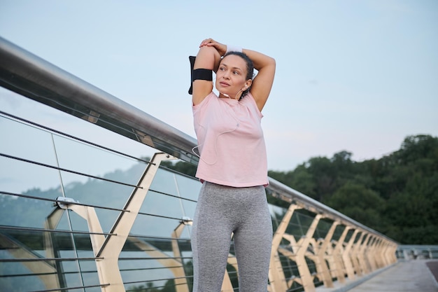 Athlétique sportive jeune femme brune étirant les bras, préparant les muscles et les ligaments, pendant l'exercice à l'extérieur avant de courir, debout à l'extérieur sur un pont de la ville sur fond de lever de soleil matinal