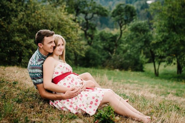 Attends bébé. famille heureuse. femme enceinte avec mari bien-aimé assis sur l'herbe, admirant le paysage. ventre rond. Le moment de tendresse sincère. fond, nature, parc, arbre, forêt, bonheur de neuf mois