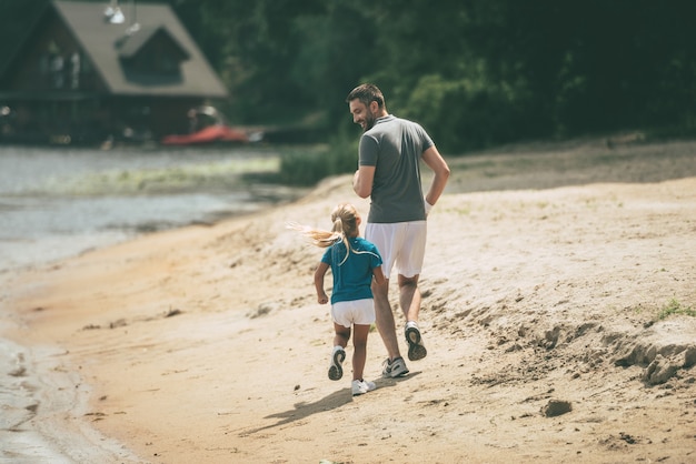 Attends-moi papa ! Vue arrière sur toute la longueur du joyeux père et fille faisant du jogging ensemble au bord de la rivière
