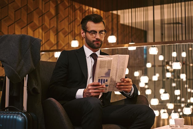 En attente d'un partenaire commercial. Jeune homme confiant lisant le journal alors qu'il était assis au restaurant de l'hôtel à proximité de la valise. Intérieur de luxe à l'arrière-plan