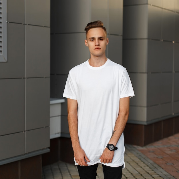 Attractive beau jeune homme dans un T-shirt élégant blanc en jean noir avec une horloge est debout dans la rue près du bâtiment moderne par une chaude journée d'été