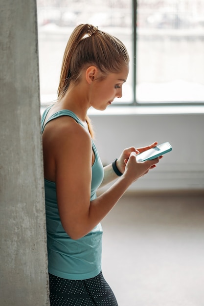 Attractive fit jeune femme vêtements de sport bleu clair faisant de la formation avec mobile au studio de loft de classe d'entraînement domestique, mode de vie sain de remise en forme