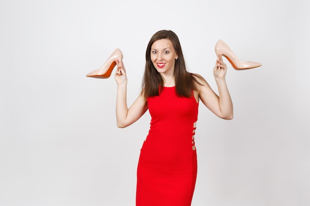 Attractive glamour caucasien à la mode jeune femme souriante aux cheveux bruns en robe rouge tenant des chaussures beiges avec la semelle rouge de ses achats isolés sur fond blanc. Copiez l'espace pour la publicité.