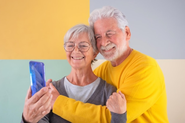 Attrayant couple de personnes âgées caucasien debout sur fond coloré isolé étreignant avec amour en regardant un téléphone intelligent