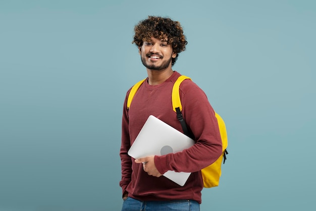 Attrayant étudiant indien souriant avec sac à dos jaune tenant un ordinateur portable, regardant la caméra isolée