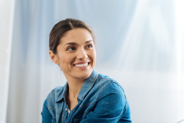 Attrayant gentil jeune femme posant sur le fond clair tout en souriant et en regardant de côté