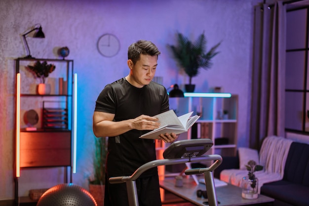Attrayant jeune homme asiatique sportif lisant un livre tout en faisant de l'exercice de fitness en cours d'exécution sur un tapis roulant