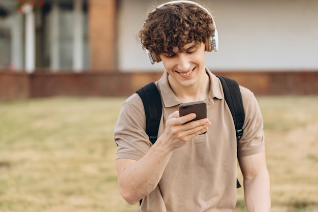 Attrayant jeune homme bouclé universitaire ou étudiant avec téléphone se promenant sur le campus