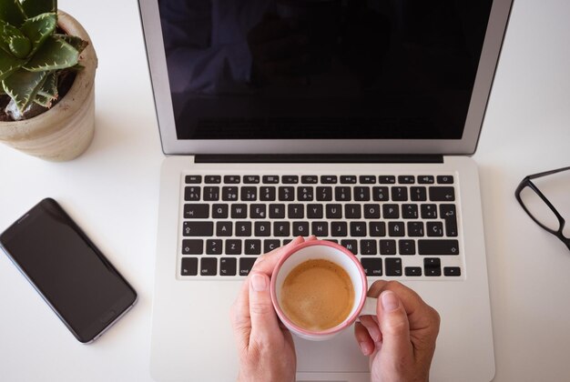 Au-dessus de la vue des mains d'une femme caucasienne tenant une tasse de café sur un bureau de clavier d'ordinateur et des appareils numériques