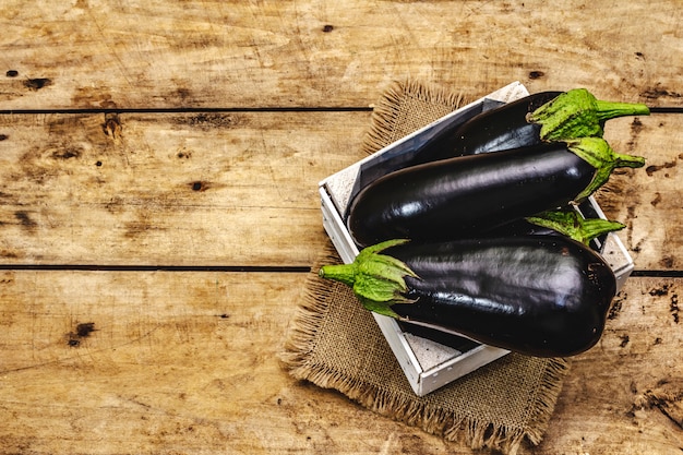 Aubergines crues entières non pelées dans une caisse en bois. Nouvelle récolte, sur un sac, fond de planches de bois, vue du dessus