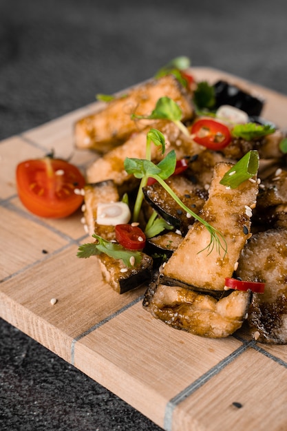 Aubergines frites sur une planche de bois, décorées de piment, ail, coriandre, graines de sésame sur une surface grise.