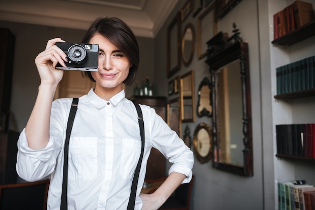 Auteure souriante en chemise blanche faisant une photo sur un appareil photo rétro avec le bras sur la hanche