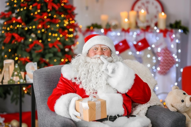 Authentique Père Noël avec boîte-cadeau assis dans un fauteuil dans une chambre décorée pour Noël