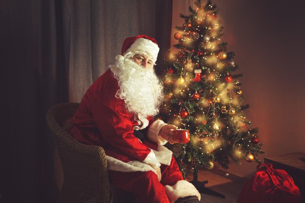 L'authentique Père Noël est assis près de l'arbre de Noël avec une tasse rouge. Intérieur de la maison. En prévision de Noël et du jour de l'An.