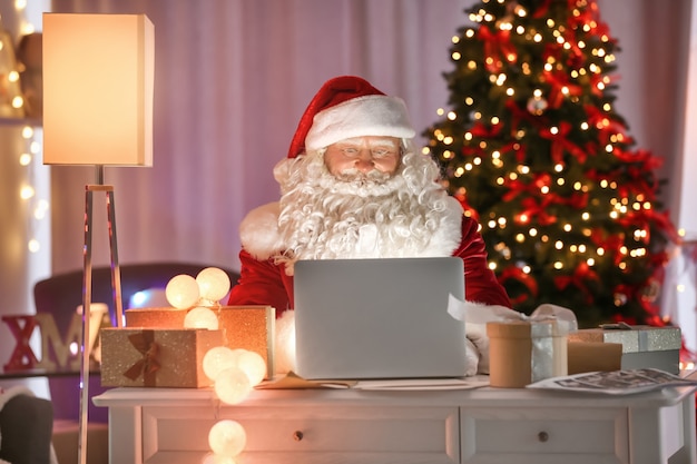 Authentique Père Noël utilisant un ordinateur portable à table dans une chambre décorée pour Noël