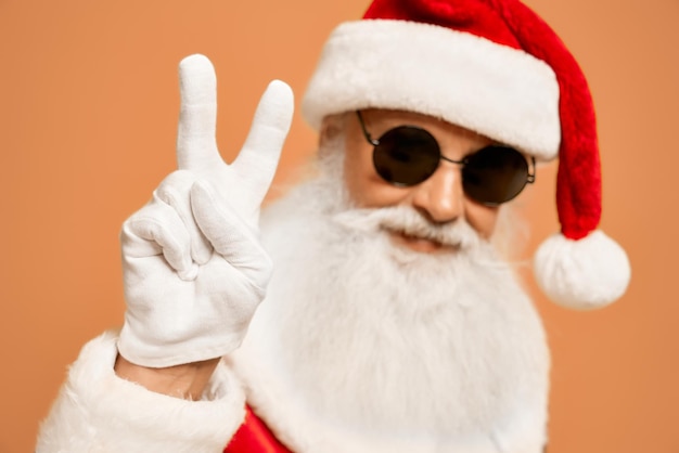 Authentique Père Noël Avec Une Vraie Barbe Grise, Portant Un Costume Traditionnel, Des Lunettes De Soleil Rondes Noires Et Des Gants Blancs Faisant Le Signe De La Victoire. Man In Santa Costume Posant Et Gesticulant En Studio.