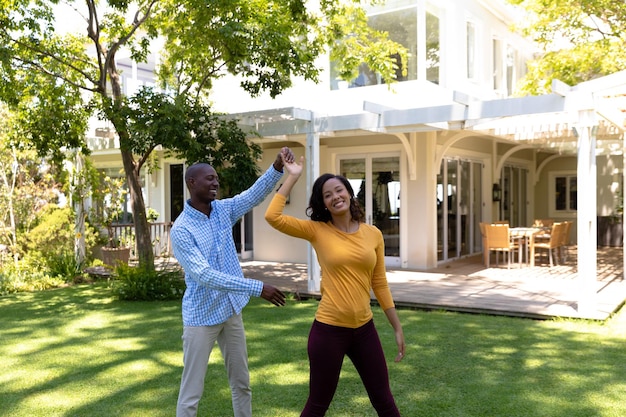 Auto-isolement en quarantaine. vue de face d'un homme afro-américain et d'une femme métisse devant leur maison dans le jardin par une journée ensoleillée, s'amusant à se tenir la main et à danser ensemble