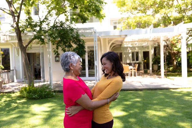 Auto-isolement en quarantaine. vue latérale d'une femme métisse et de sa mère aînée debout devant leur maison dans le jardin par une journée ensoleillée, s'embrassant, se regardant et souriant