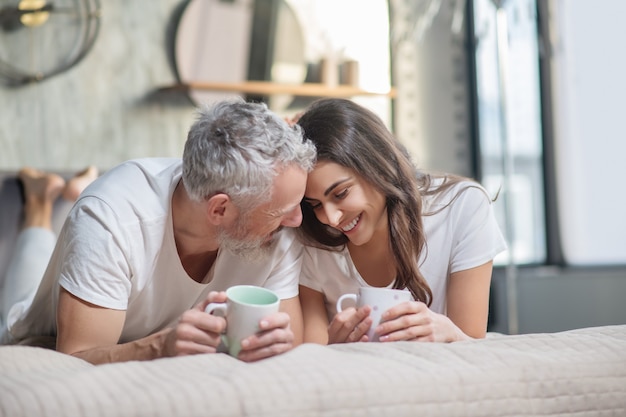 Autochtones. Sourire heureux homme et femme avec des tasses allongé sur le lit sur l'estomac touchant les têtes