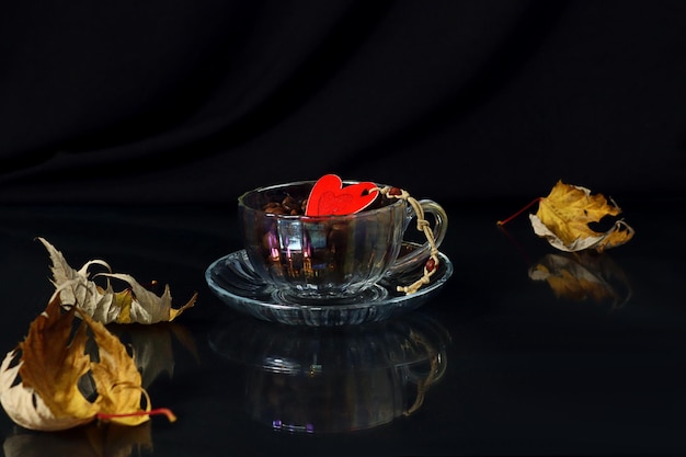 Automne confortable une tasse avec des grains de café un coeur décoratif rouge dedans des feuilles d'érable sèches à proximité réflexion des objets fond sombre espace pour le texte