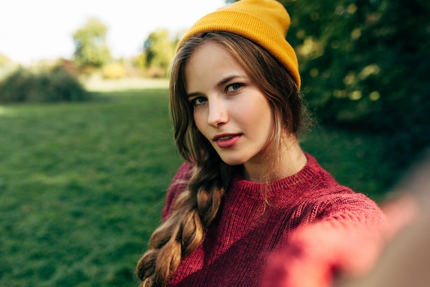 Autoportrait de la belle jeune femme blonde souriante et regardant vers la caméra portant un pull et un chapeau posant sur fond de nature dans le parc Jolie femme prenant selfie Les gens voyagent style de vie