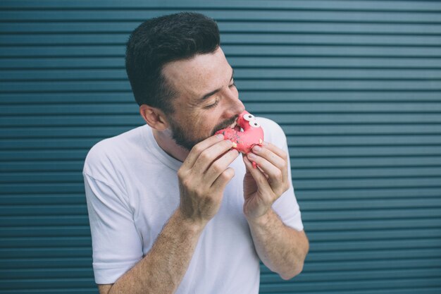 Une autre photo d'un homme mordant un morceau de beignet rouge. Il aime les bonbons. Isolé sur rayé