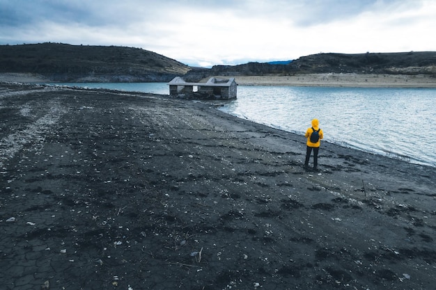 Aventurier routard anonyme avec veste jaune regardant une maison en ruine et abandonnée près du lac