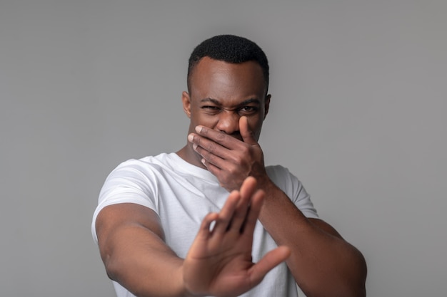 Aversion, émotion. Jeune adulte homme à la peau sombre avec grimace couvrant sa bouche avec des gestes de la main en repoussant en studio photo