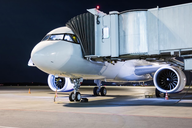 Un avion de ligne blanc se tient à la passerelle d'embarquement connecté à une alimentation externe sur un tablier de nuit de l'aéroport