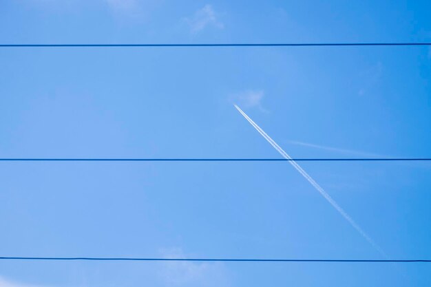 Un avion de ligne et sa traînée de fumée blanche derrière des fils à haute tension contre le ciel bleu