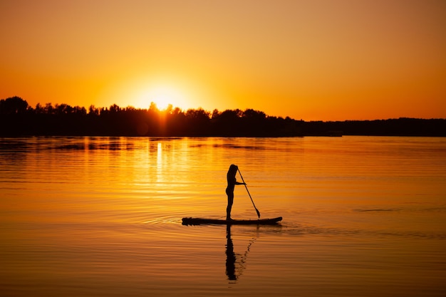 Aviron sur planche sup femme avec rame dans les mains avec sa réflexion sur l'eau sur un lac calme avec coucher de soleil apaisant et arbres en arrière-plan Mode de vie actif