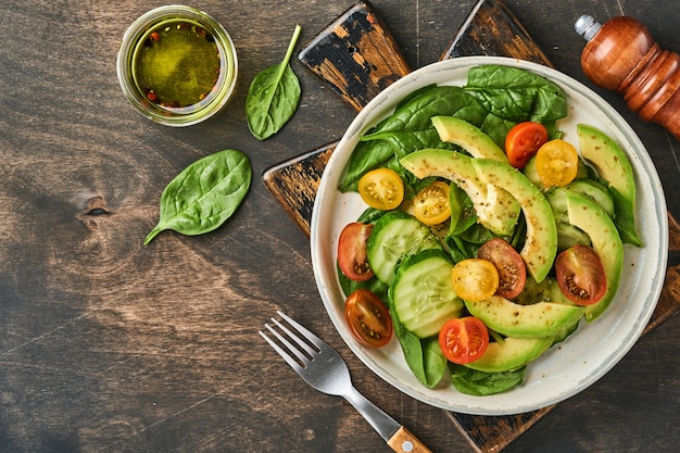 Avocat, rouge, jaune, tomate cerise noire, épinards et salade fraîche de concombre avec des épices, du poivre et de l'huile d'olive dans un bol gris sur fond de table en bois ancien. Concept d'alimentation saine. Vue de dessus.