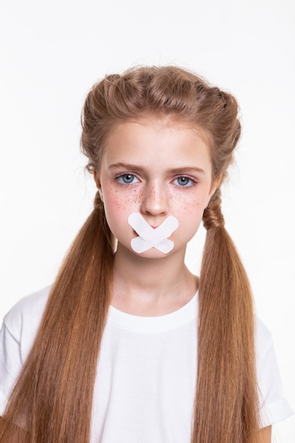 Avoir la bouche couverte. Jolie petite fille aux cheveux longs en t-shirt blanc triste à cause de la bouche fermée