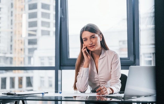 Avoir une conversation par téléphone Jeune femme en vêtements formels blancs est à l'intérieur dans le bureau moderne